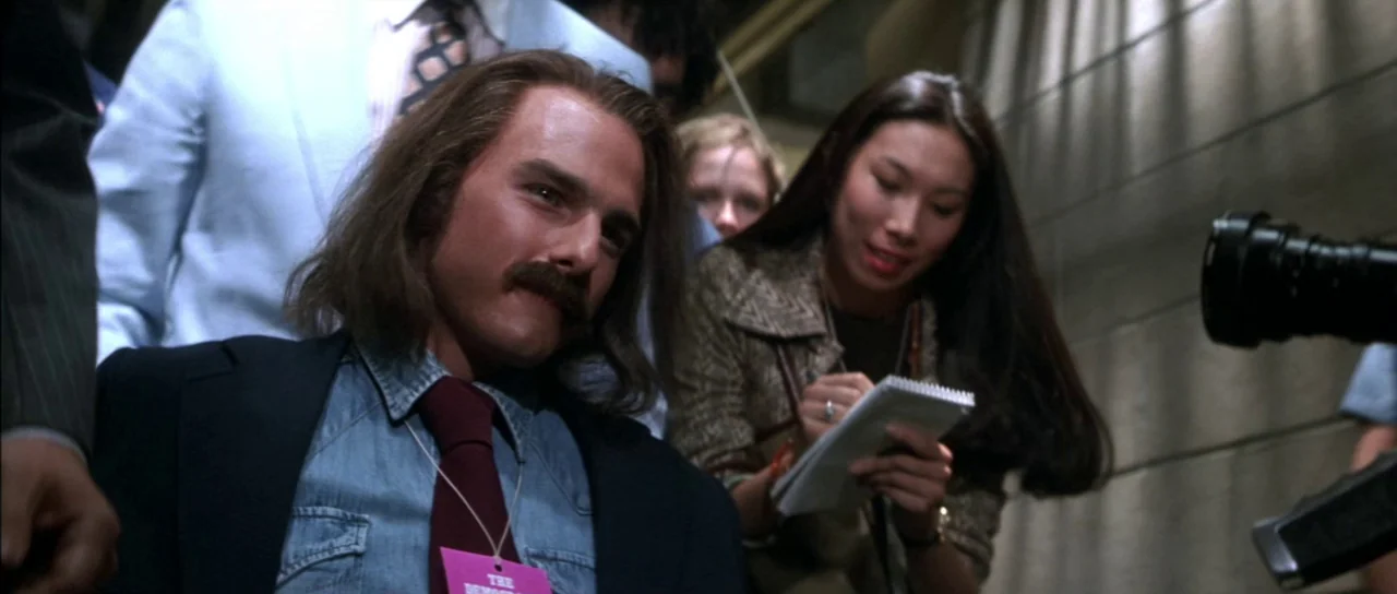Born On The Fourth of July (1989) movie still: medium — An interior hallway with beige tiled walls and a crowd of people nearby. A man with a…
