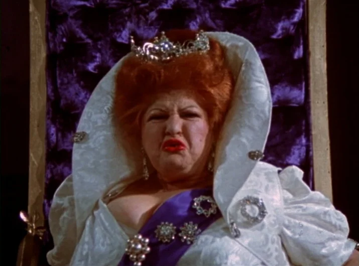 Desperate Living (1977) movie still: closeup — A woman sits on a throne against a dark purple tufted velvet backdrop. She wears a crown and formal…