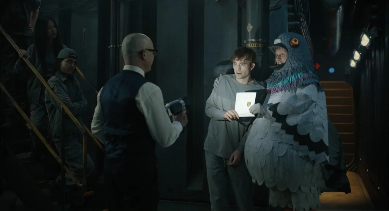 Mickey 17 (2025) movie still: medium — An industrial metallic corridor with dark walls and exposed pipes on the sides. A man in a bird costume and a…