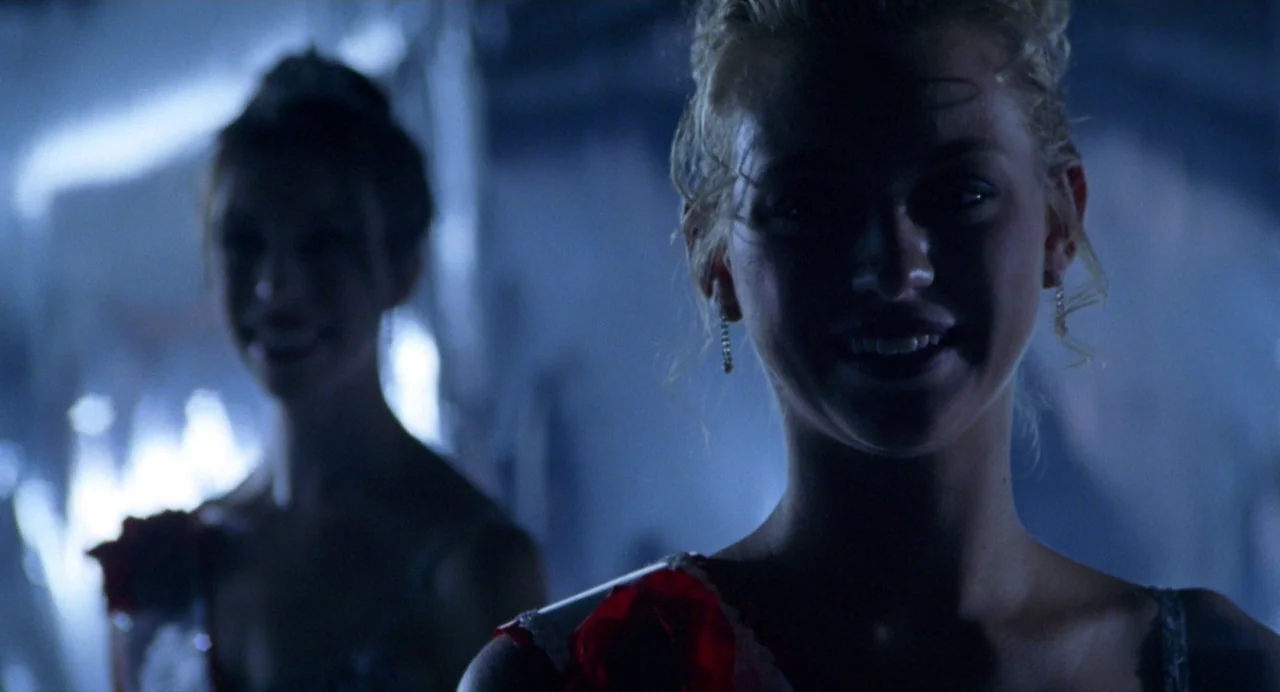 Drop Dead Gorgeous (1999) movie still: medium — Two young women with updos and corsages stand in a dark, dimly lit gymnasium. They are both smiling…