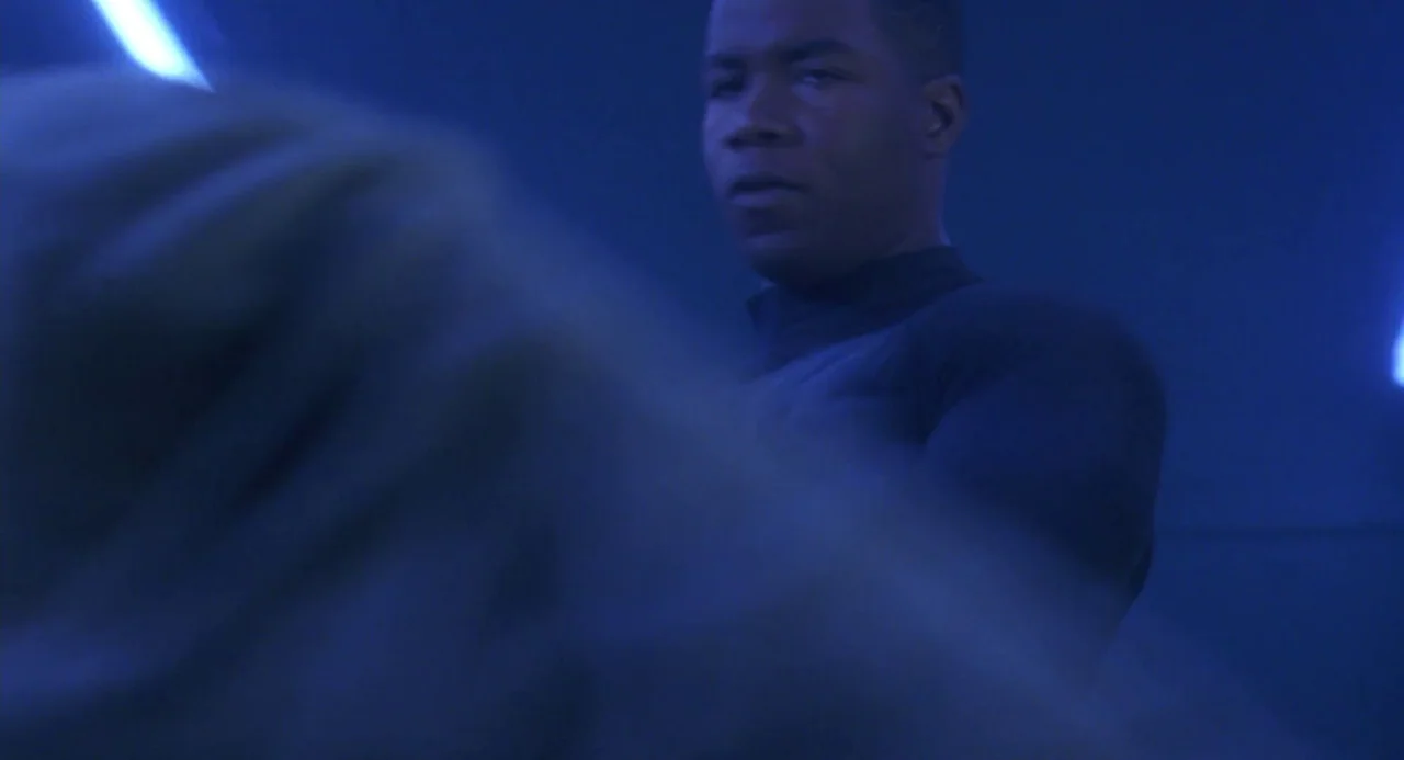 Universal Soldier: The Return (1999) movie still: medium — A dimly lit room bathed in intense blue light. A young man stands, holding a glowing blue…