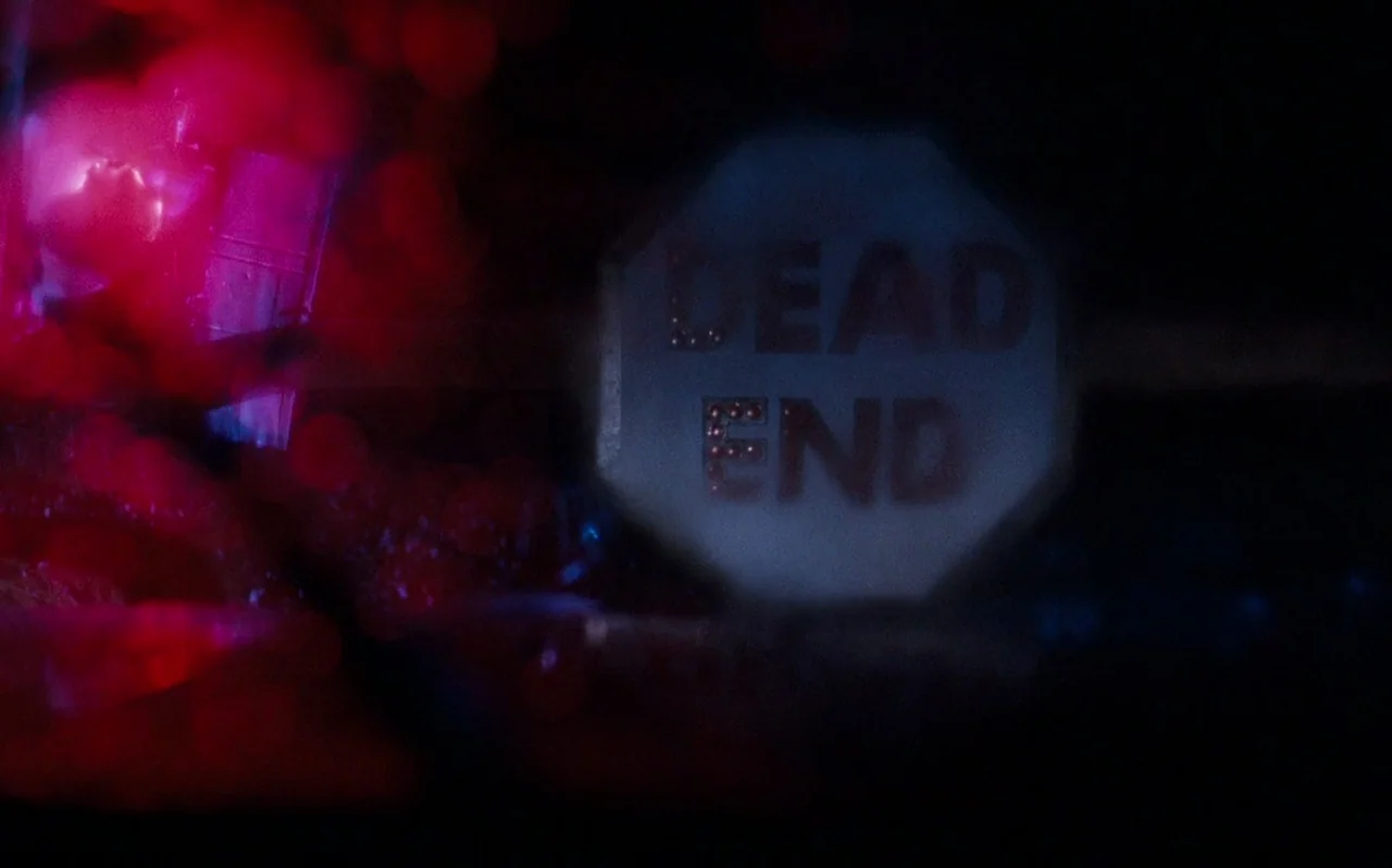 The Rocky Horror Picture Show (1975) movie still: closeup — A reflective traffic sign reads 'DEAD END' in the darkness of the night. Flashing red…