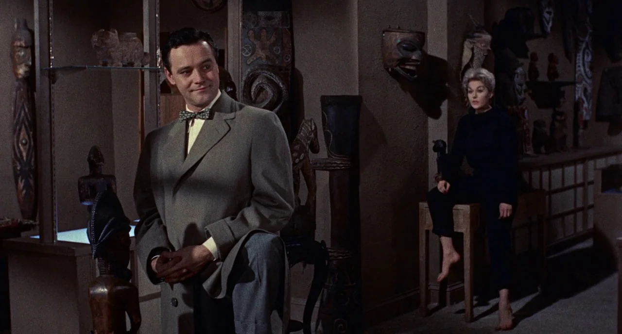 Bell Book and Candle (1958) movie still: medium — A dimly lit shop filled with assorted wooden carvings and ethnographic wall masks. A man stands in…