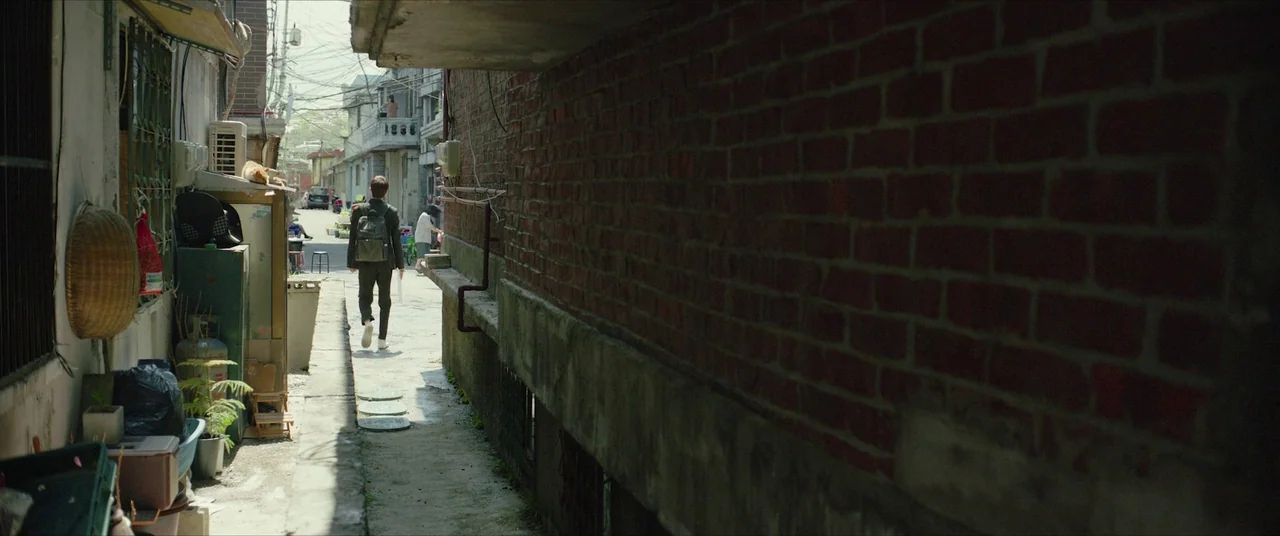 Parasite (2019) movie still: fullbody — A narrow alleyway features a tall red brick wall and various cluttered household items. A man wearing a…