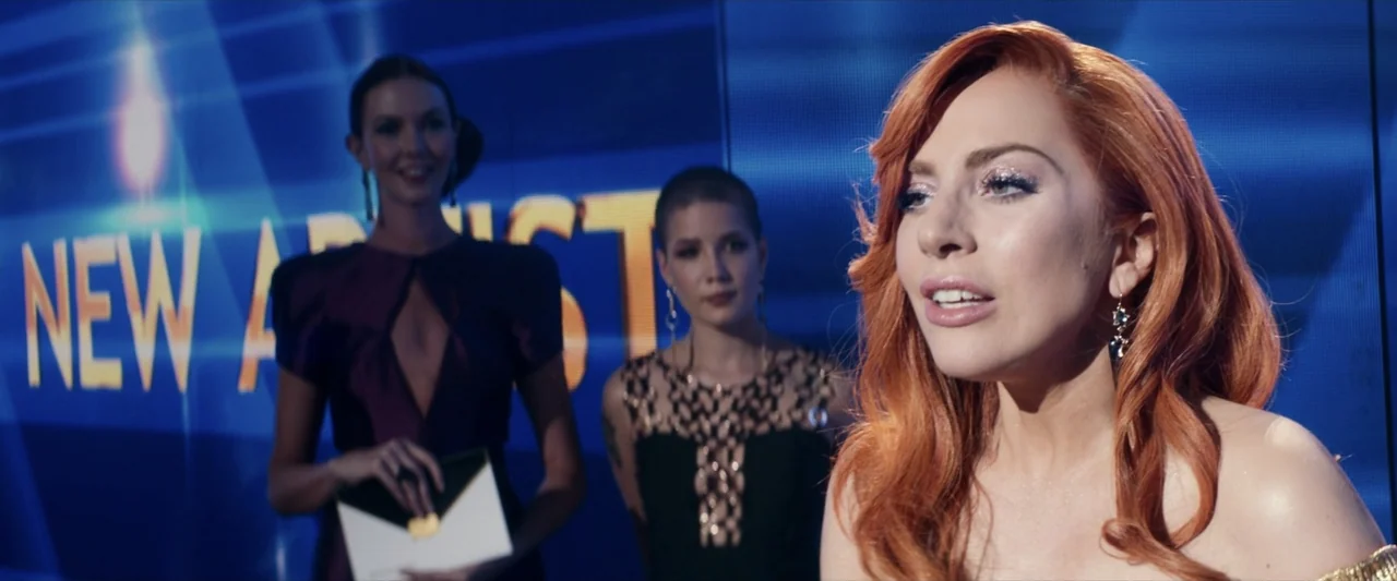 A Star Is Born (2018) movie still: medium — An brightly lit award ceremony stage features a large blue digital display background. A woman with…