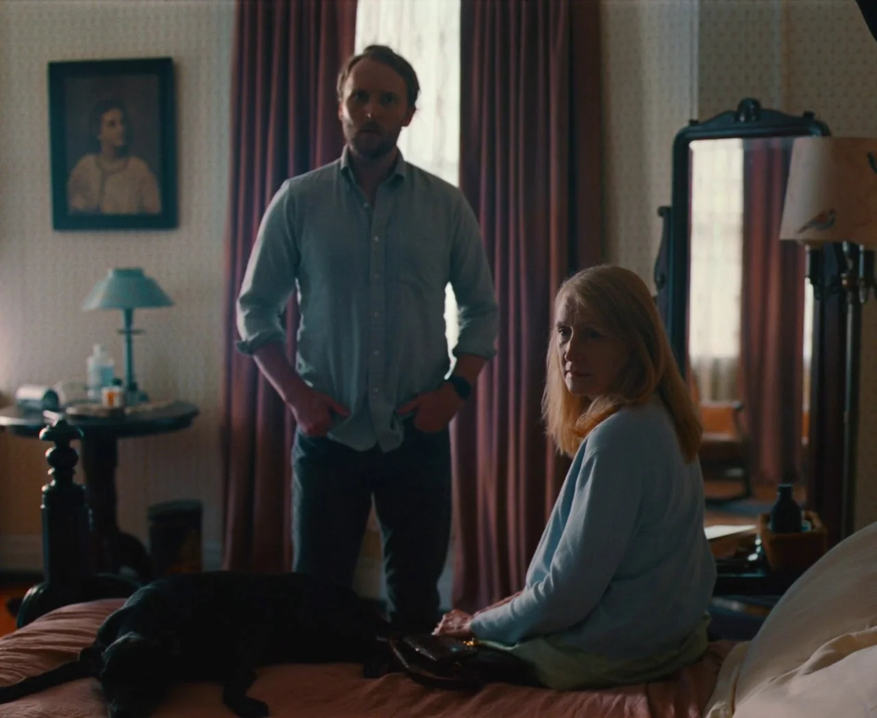 Monica (2022) movie still: medium — A furnished bedroom with patterned wallpaper features a bed, a framed portrait, and a mirror. A man stands…