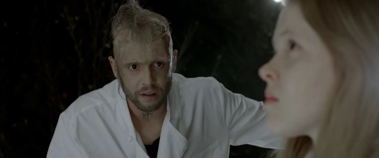 The Lure (2015) movie still: medium — A dark outdoor setting at night features blurred dark foliage in the background. A man with a neck tattoo in a…