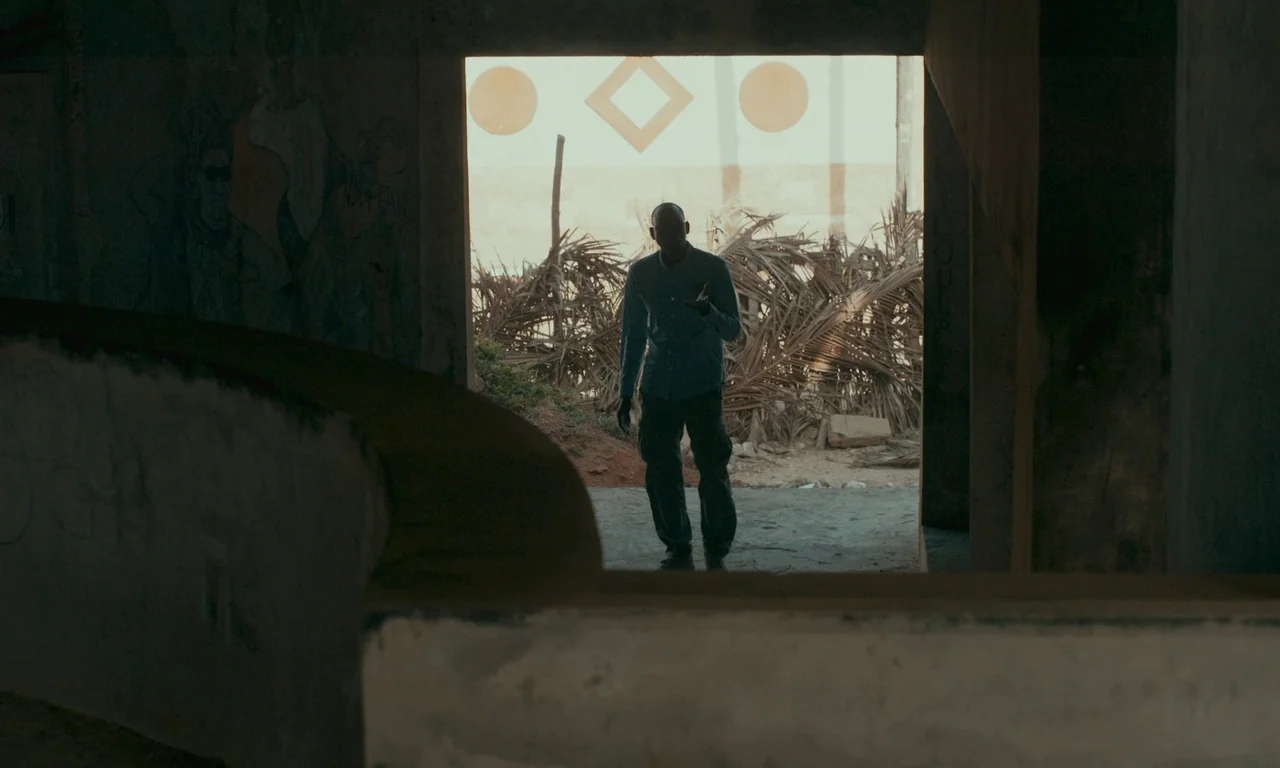 Atlantics (2019) movie still: fullbody — A dark, dilapidated interior space looks out onto a sun-drenched outdoor area with dried palm leaves. A man…