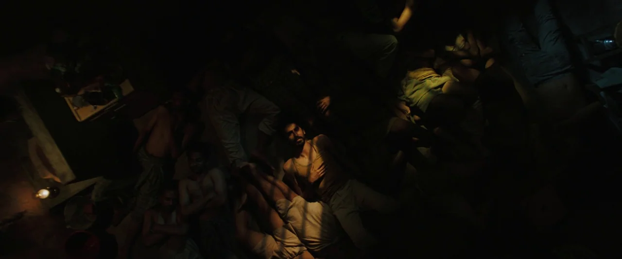 Monkey Man (2024) movie still: wide — A dark dormitory floor is densely packed with multiple men lying side by side. The central figure lies awake…