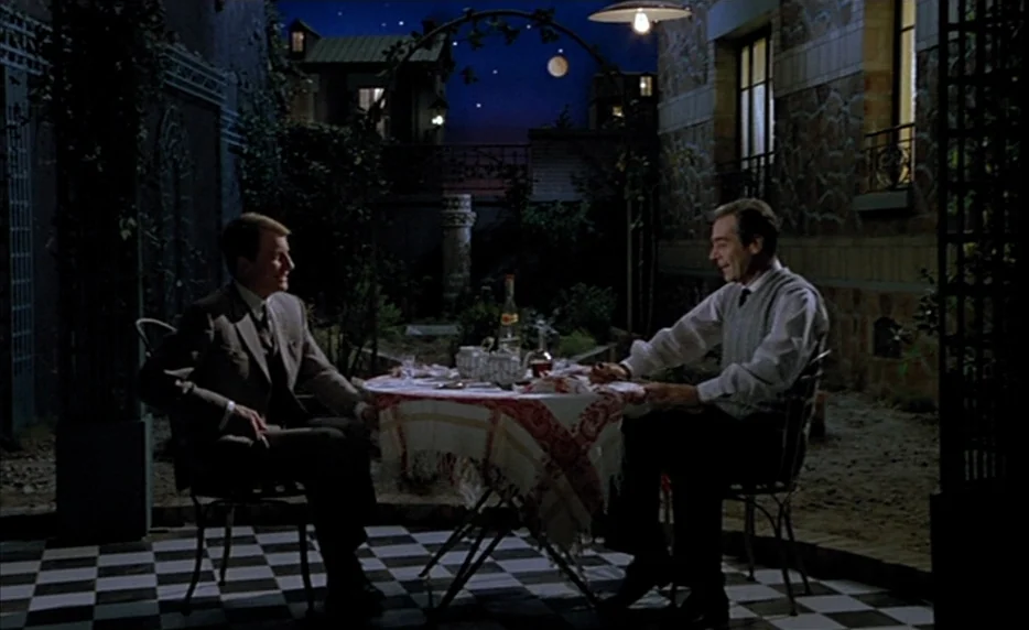 Mélo (1986) movie still: wide — A patio with a checkered floor sits under a clear night sky. Two men sit at a small table covered by a decorative…