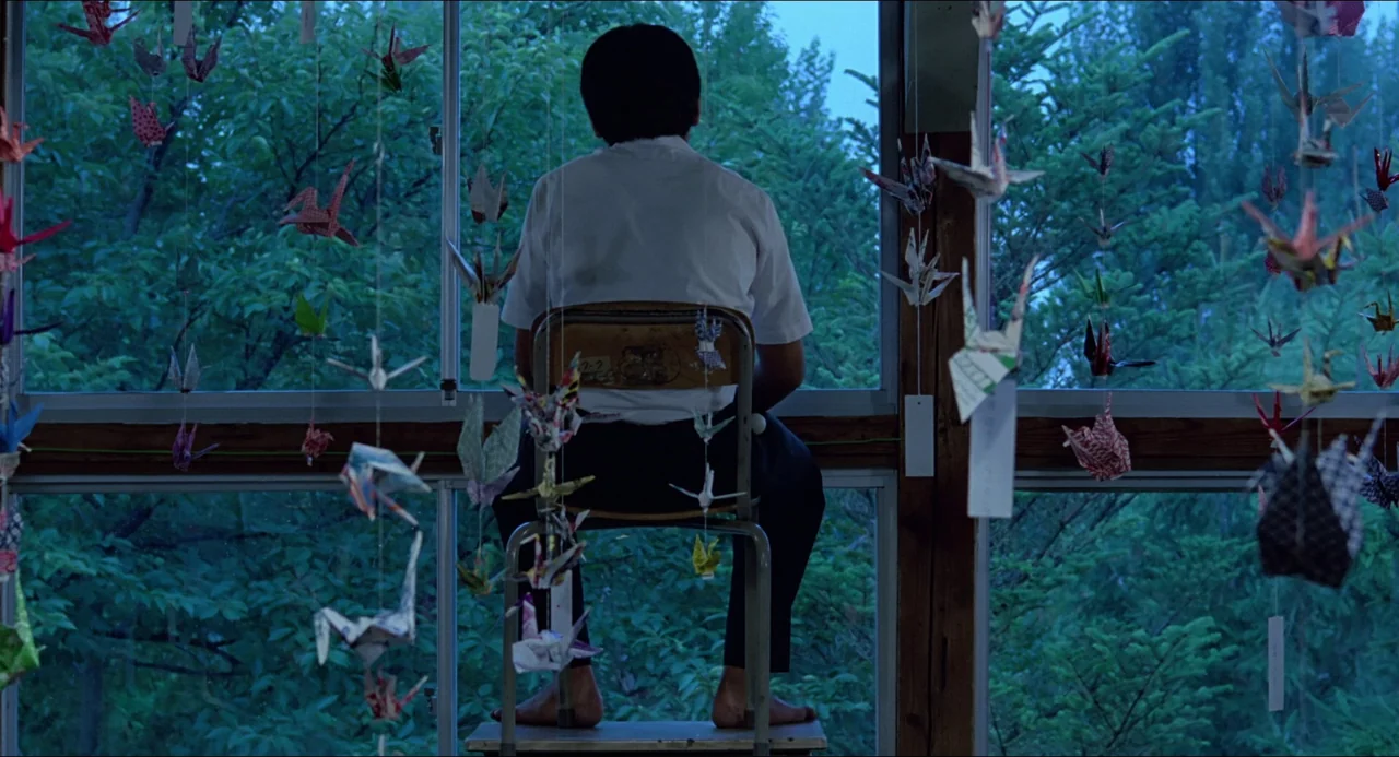 Typhoon Club (1985) movie still: fullbody — A student sits in an old classroom facing a window covered in hanging paper cranes. Outside the window,…