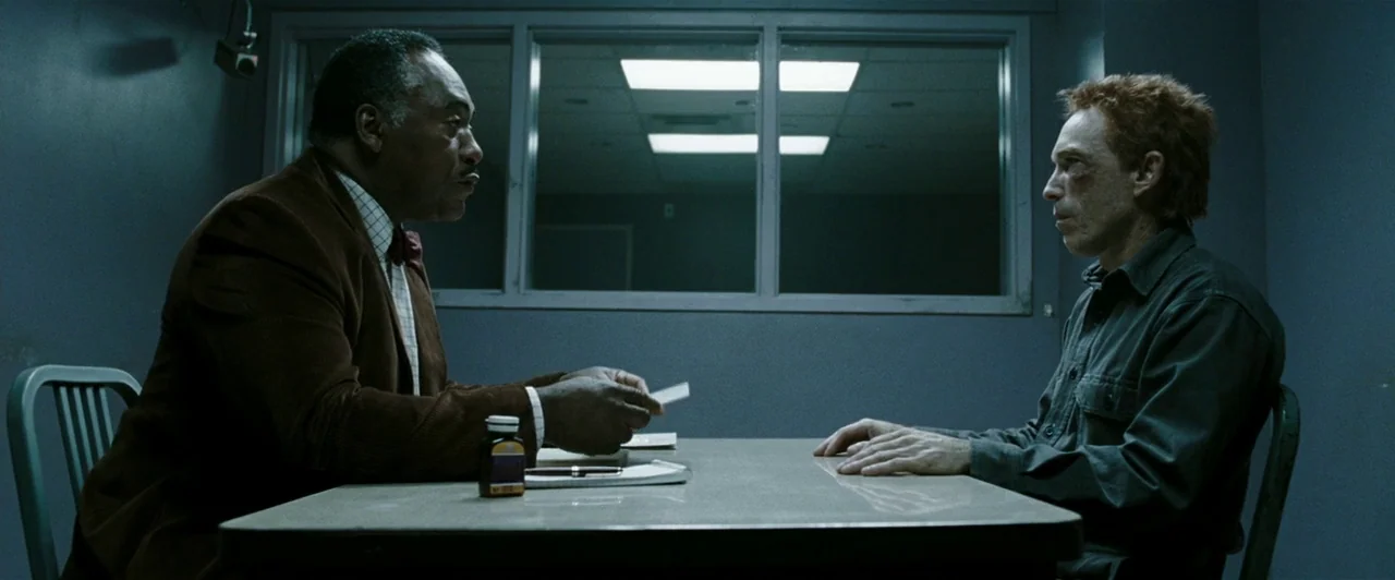 Watchmen (2009) movie still: medium — A stark room features blue-grey walls and a single metal table for interrogation. Two men sit directly across…