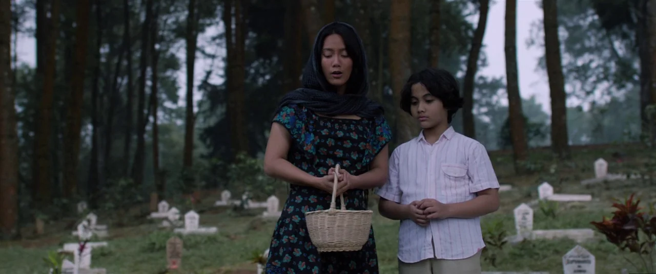 Satan’s Slaves (2017) movie still: medium — A woman and a young boy stand in a hillside cemetery with numerous markers. The woman holds a small…