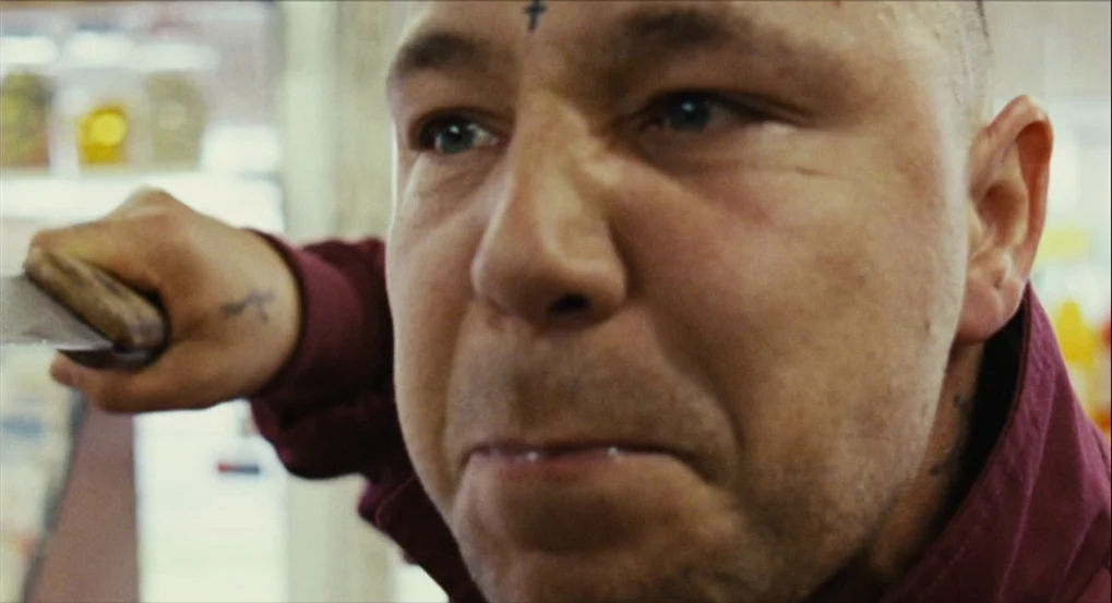 This Is England (2006) movie still: closeup — A man stands inside a brightly lit retail space with shelves visible in background. He clenches his…