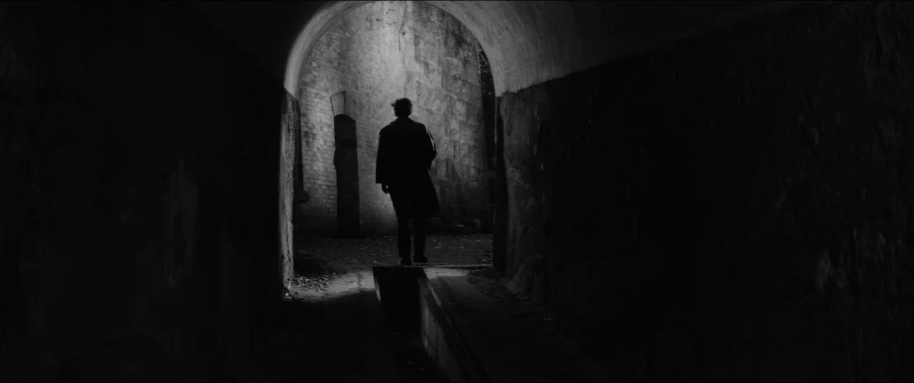 Branded To Kill (1967) movie still: fullbody — An arched stone tunnel leads toward an opening into a bright courtyard area. A solitary man in a long…