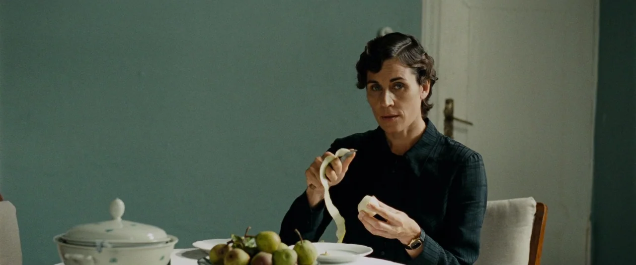 Phoenix (2014) movie still: medium — A quiet dining room with muted teal walls and simple 1940s-style furniture. A woman sits at a white-clothed…