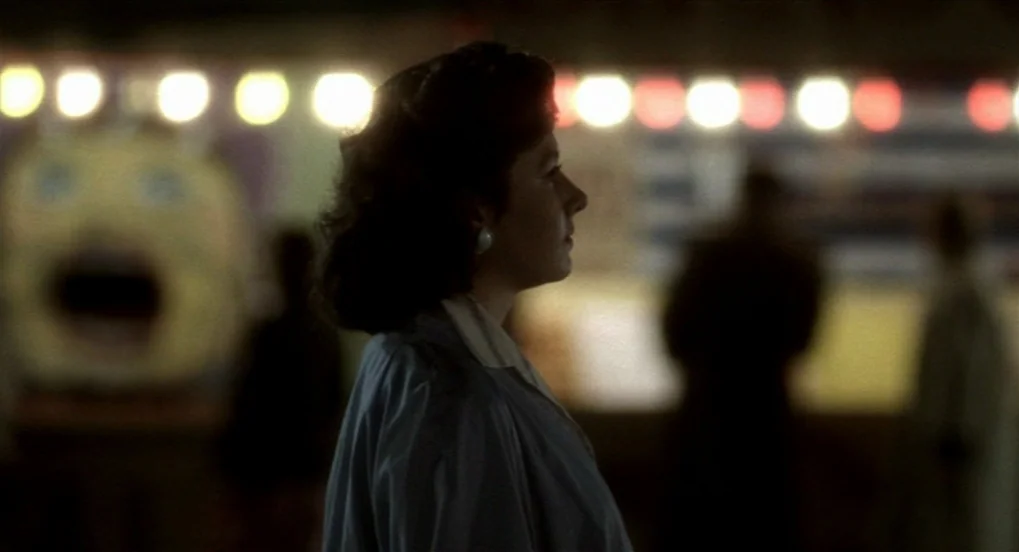 The Long Day Closes (1992) movie still: medium — An outdoor carnival area at night with out-of-focus fairground lights in the background. A woman in…