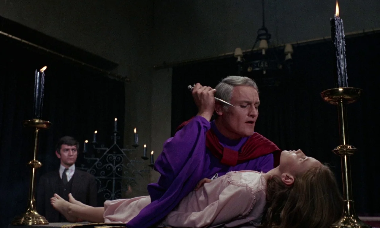 The Devil Rides Out (1968) movie still: medium — A dimly lit room with black drapes serves as a ritual space for two individuals. An older man in…