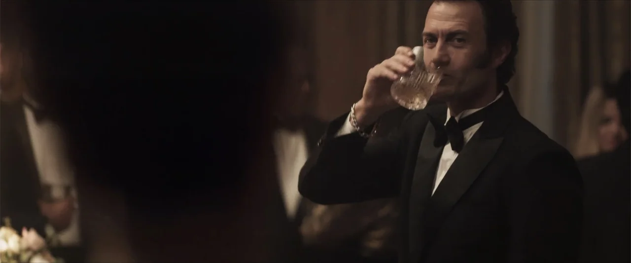 Angela (2023) movie still: medium — A crowded dimly lit ballroom features guests wearing formal black-tie evening attire. A man in a tuxedo stands…