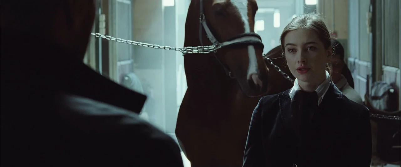 Anna (2013) movie still: medium — An indoor equestrian facility features wooden stalls and a bay horse in the background. A young woman in a riding…