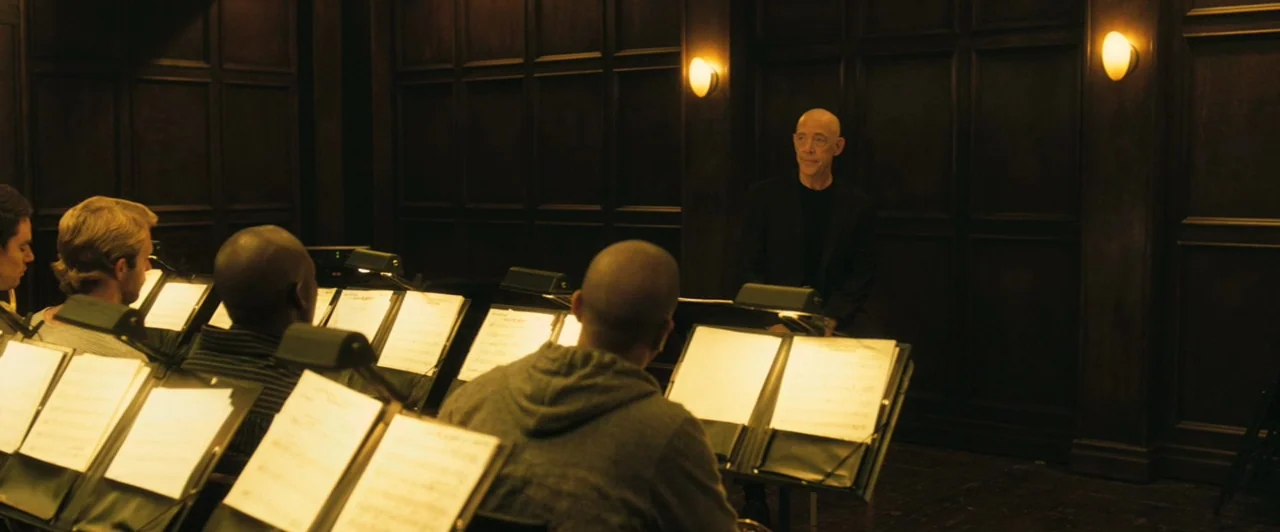 Whiplash (2014) movie still: wide — A dark, wood-paneled room contains several musicians seated behind music stands. A bald older man sits in a…