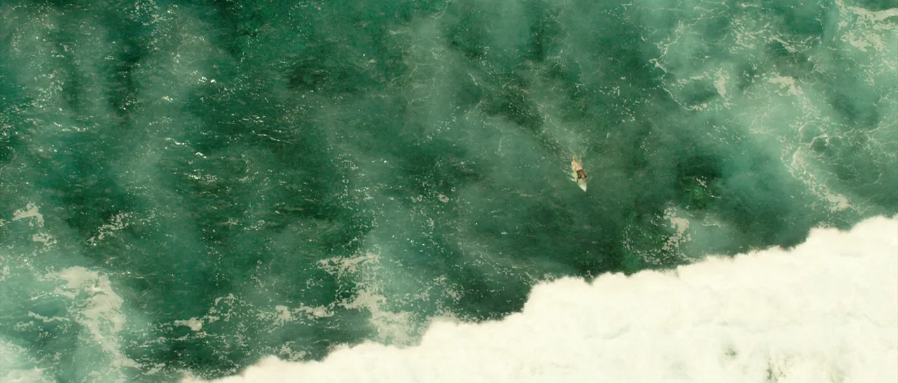 The Shallows (2016) movie still: wide — A vast expanse of deep green ocean water is broken by white turbulent surf. A lone surfer paddles on a board…