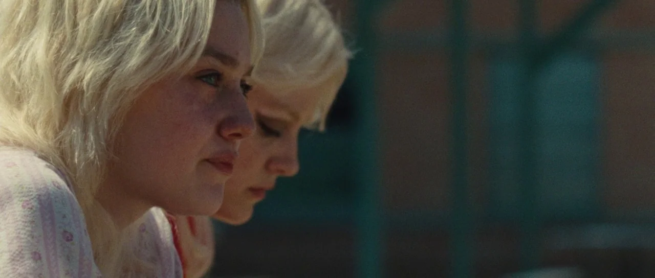 The Runaways (2010) movie still: closeup — Two young women with blonde hair sit together in a bright outdoor setting. They both gaze forward with…