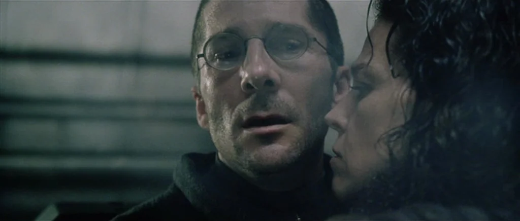 Alien: Resurrection (1997) movie still: closeup — Two individuals stand in a dimly lit, nondescript industrial setting with horizontal structural…