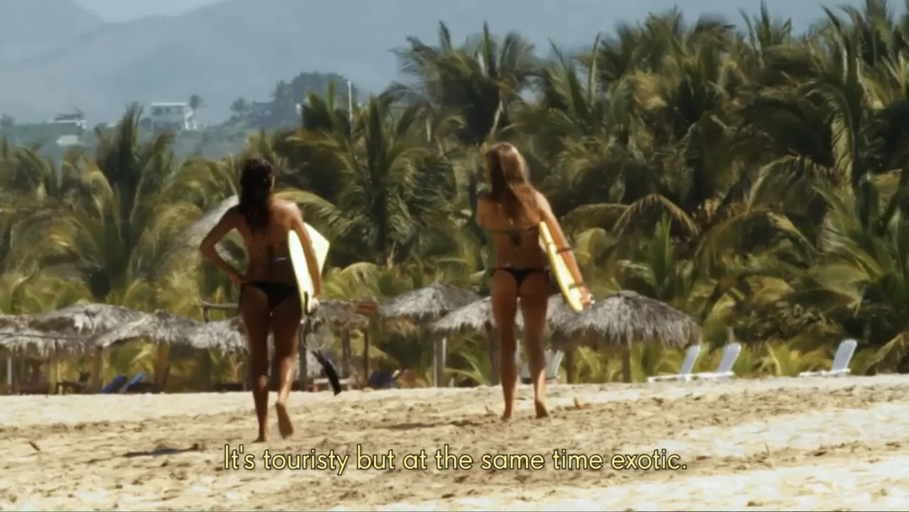 El sueño (2020) movie still: fullbody — A sandy beach lined with numerous palm trees and small thatched-roof structures. Two women in bikinis walk…