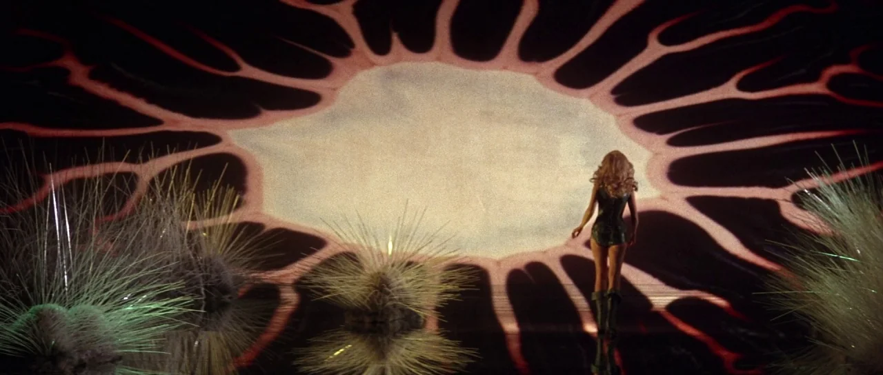 Barbarella (1968) movie still: wide — An abstract, dark environment features a large, glowing, organic-shaped light source on a wall. A woman walks…