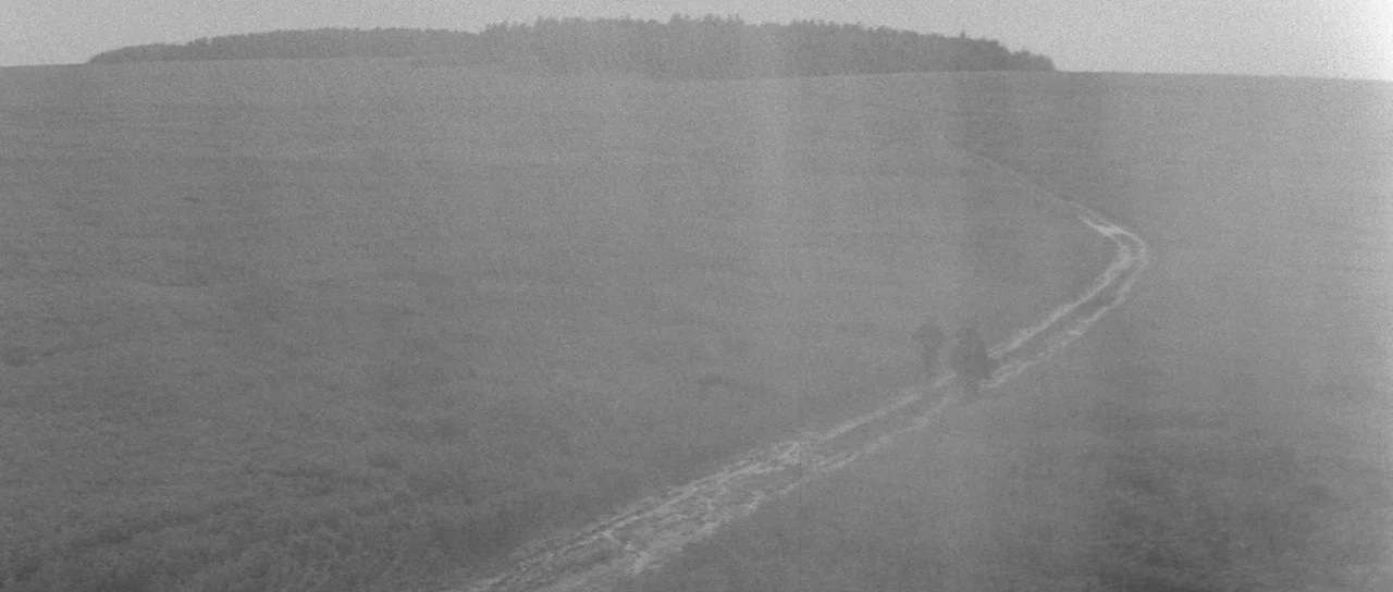 Andrei Rublev (1966) movie still: wide — A vast, open field is covered in heavy fog under a flat, grey sky. Two small, indistinct figures walk…