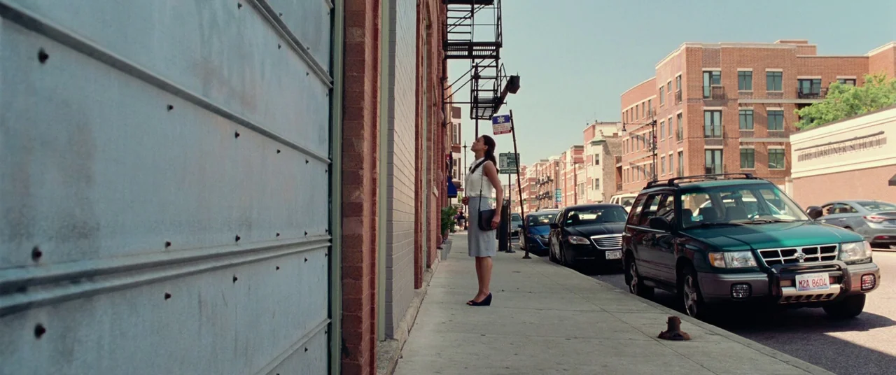 Widows (2018) movie still: wide — An urban sidewalk runs alongside a tall brick building with a metal garage door. A woman stands in profile against…