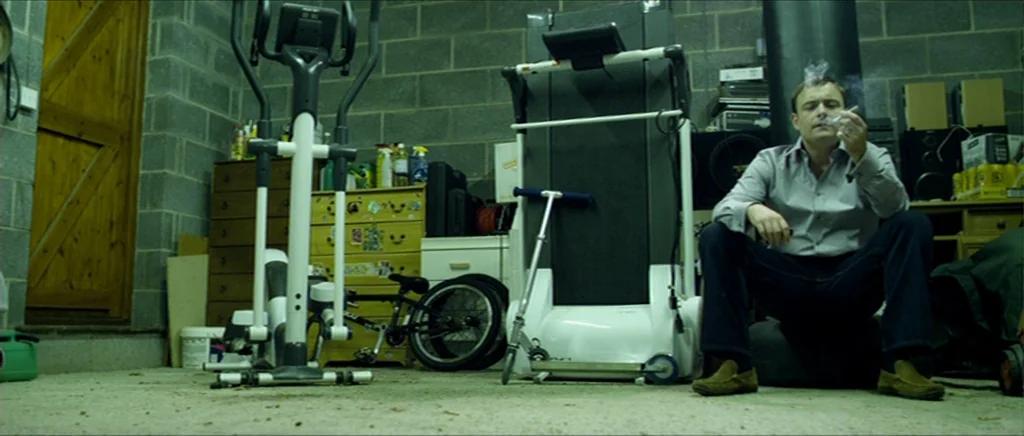Kill List movie still: fullbody — A basement room features grey concrete block walls and assorted gym equipment. A man sits alone on a low dark…