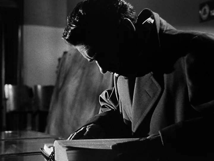 Nightmare Alley (1947) movie still: medium — A man in a heavy coat examines a large book inside a dark building. He leans over the page with his…
