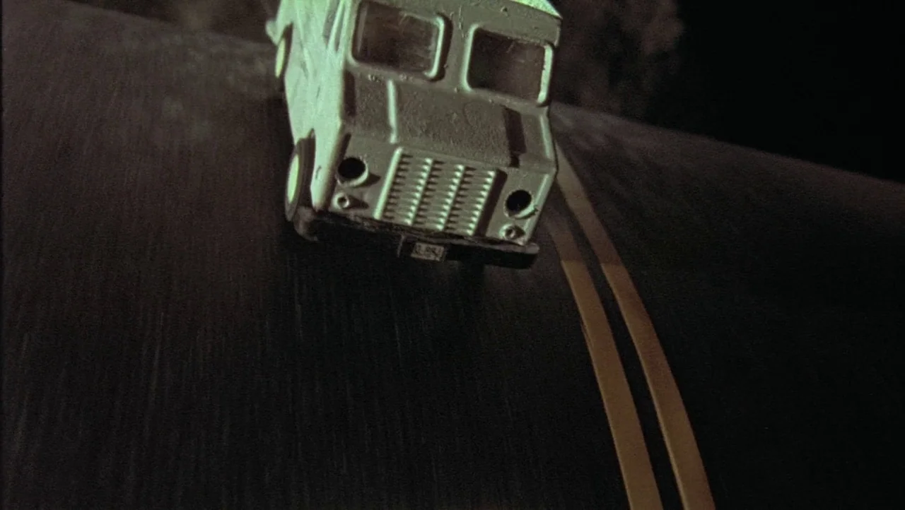 Hard Core Logo movie still: closeup — A miniature metal truck toy is positioned on a dark asphalt road at night. Two solid yellow lines run parallel…