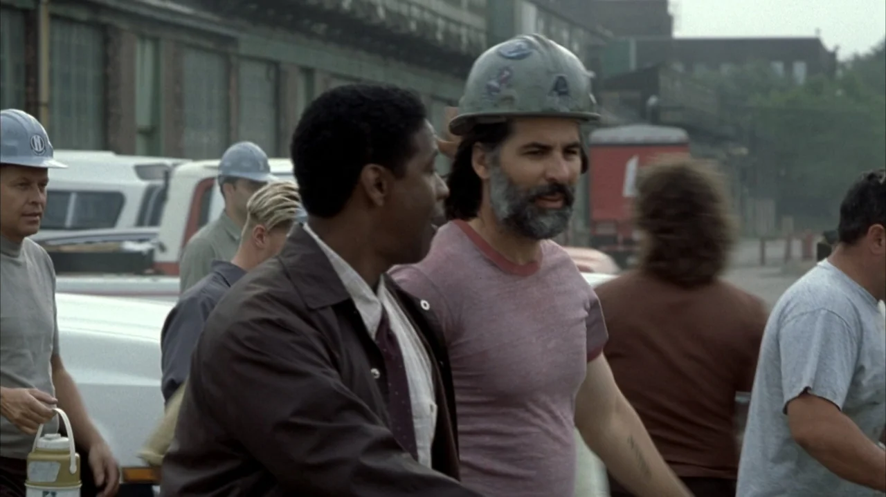 John Q (2002) movie still: medium — An industrial parking lot features several white work trucks and a large factory building. A man in a dark…