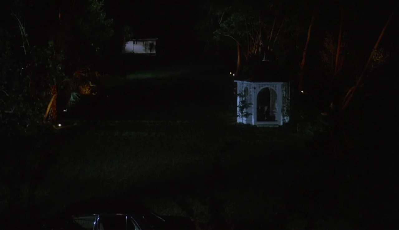 Body Heat (1981) movie still: establishing — A dark, grassy lawn at night features a lone, brightly lit white gazebo. A distant building is visible…