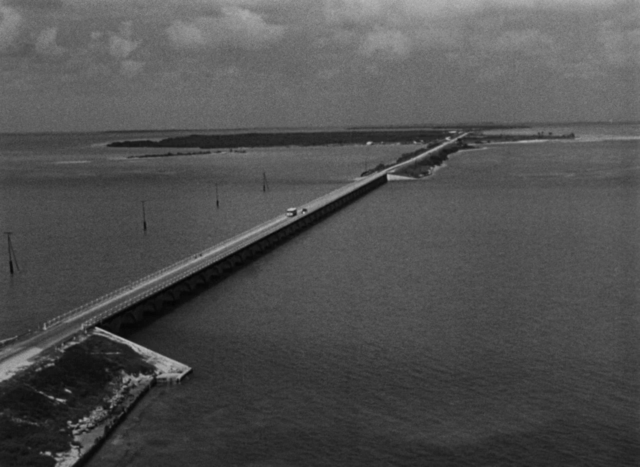 Key Largo (1948) movie still: establishing — A long concrete bridge stretches across a wide expanse of ocean water under clouds. A small vehicle…