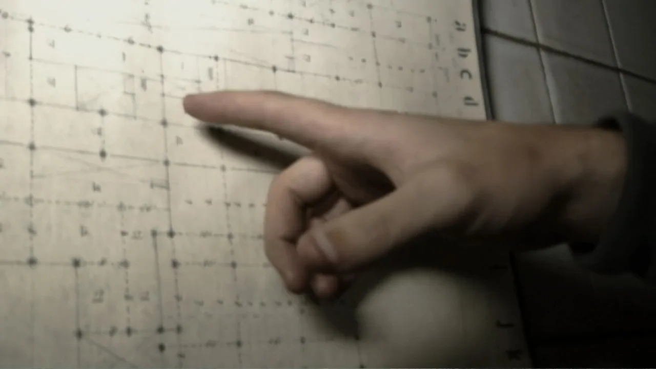 Lost (2010) movie still: closeup — A close-up view shows a hand resting against a detailed grid-based technical blueprint. The person is pointing…