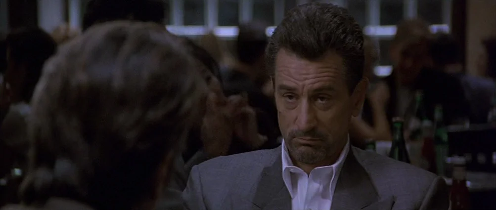 Heat (1995) movie still: medium — A dimly lit restaurant setting features blurred patrons in the background behind a man. The man sits at a table…