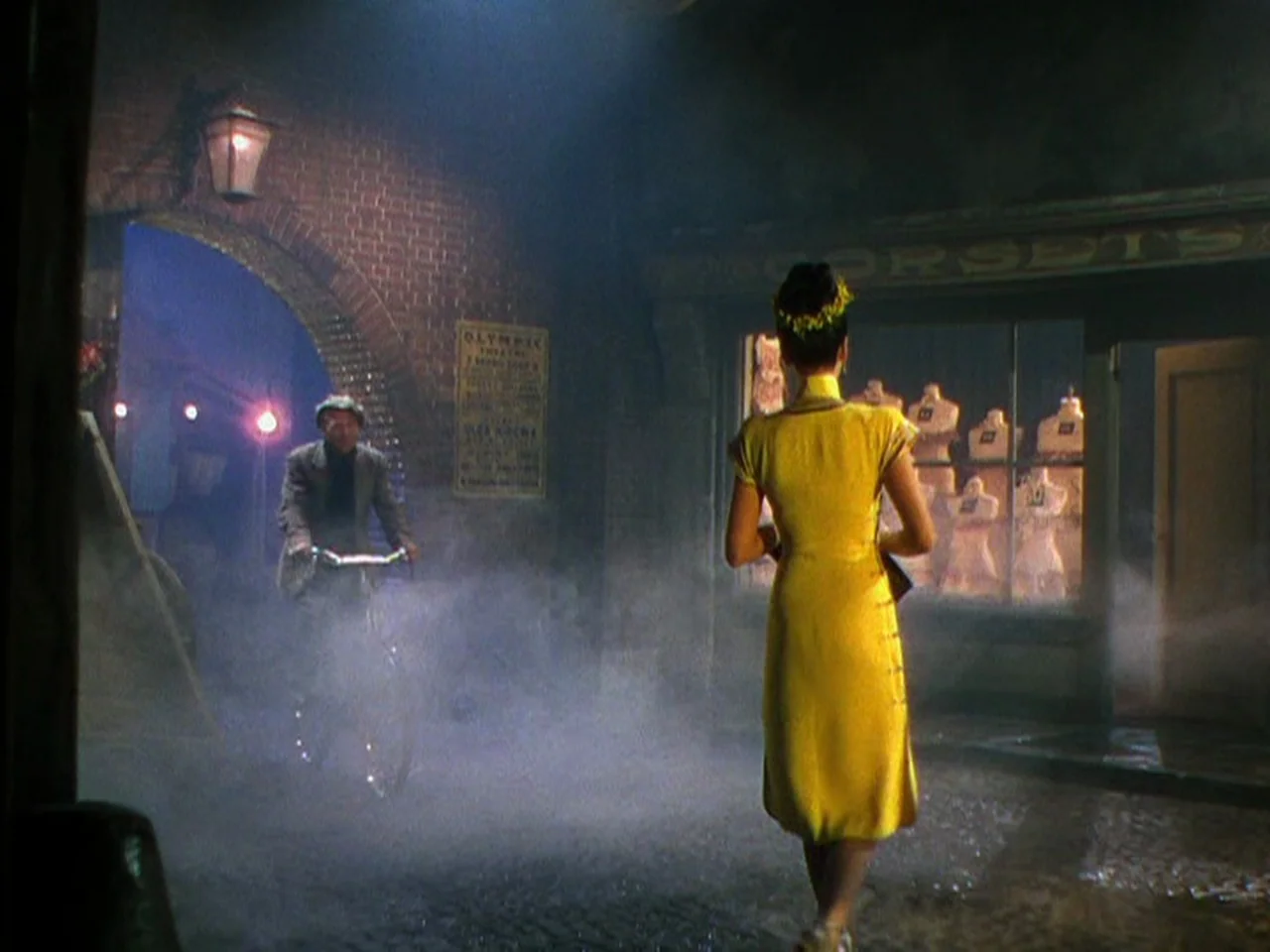 Ziegfeld Follies (1945) movie still: wide — A wet cobblestone street at night features thick ground fog and a shop window. A man rides a bicycle…