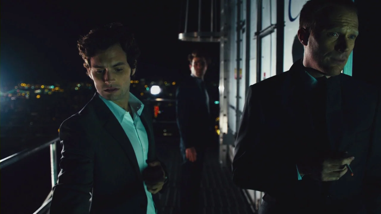Margin Call (2011) movie still: medium — A dark, narrow metal walkway at the top of a skyscraper overlooks a city. Two men stand in the shadows on…