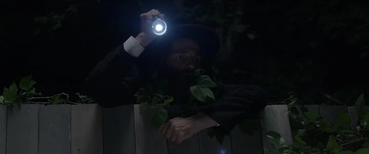 To Dust - Nightmares (2018) movie still: medium — A man in a dark suit peers over a wooden fence at night. He holds a bright flashlight that…
