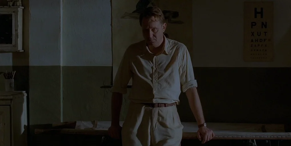 Dominion: Prequel to the Exorcist (2005) movie still: medium — A dimly lit room contains an examination table and an eye chart on the wall. A man in…