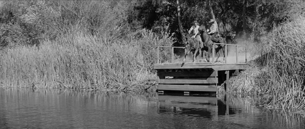 Love Me Tender movie still: wide — Two men on horseback cross a simple wooden bridge extending over a calm river. Tall reeds and dense foliage…