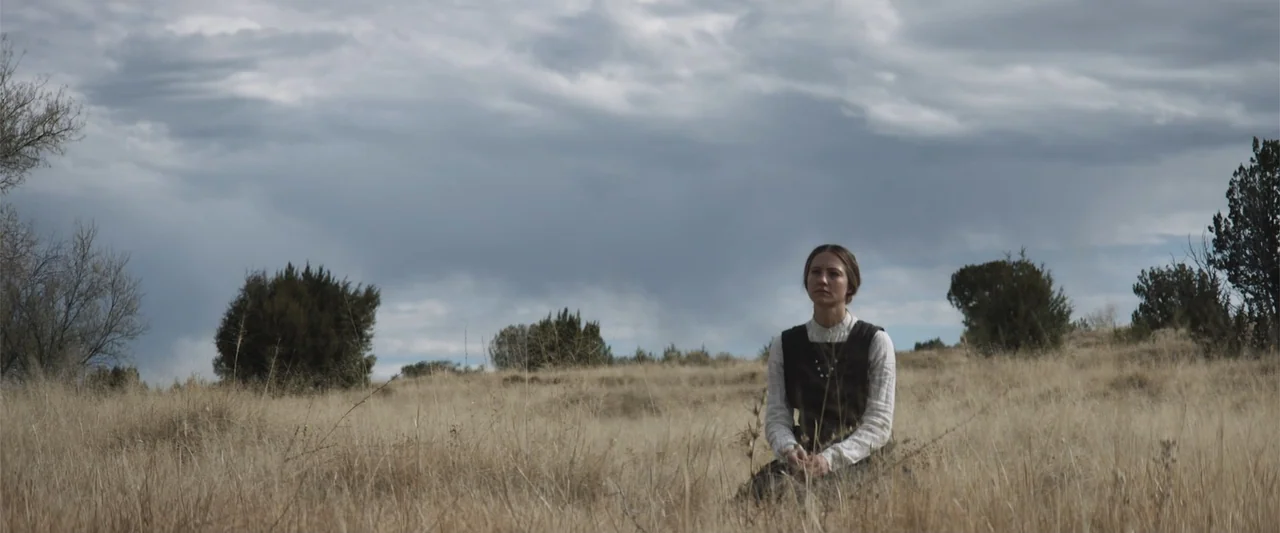 The Wind (2005) movie still: medium — A woman kneels in a dry, open field under a cloudy, overcast sky. She rests in the tall grass while facing…
