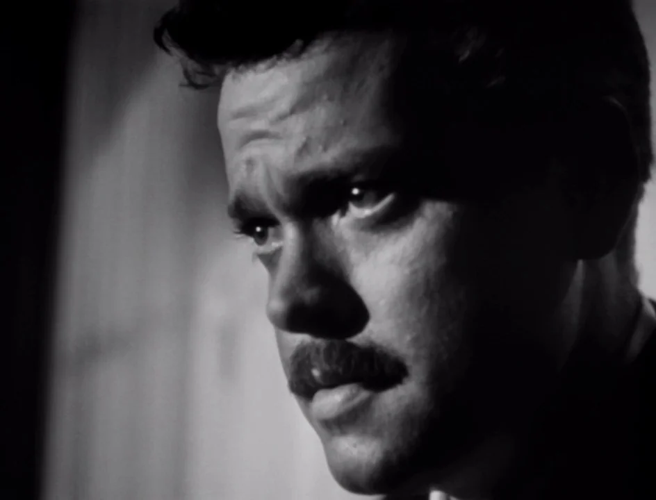 The Stranger (1946) movie still: closeup — A dark, minimally lit space with soft texture on the wall behind the subject. A man with a mustache…