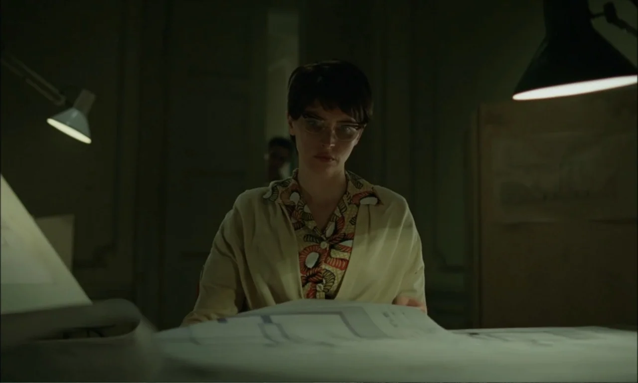 The Brutalist (2024) movie still: medium — A dim office workspace features blueprints spread across a table with two desk lamps. A young woman with…