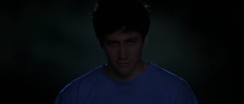 Donnie Darko movie still: closeup — A man stands in extreme darkness with only his face and shoulders illuminated. He stares forward with a subtle…