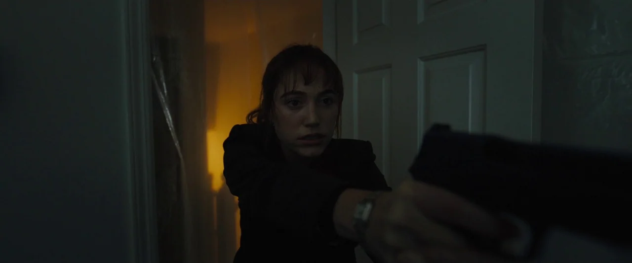Longlegs (2024) movie still: medium — A dimly lit indoor passage featuring a white door and a plastic-covered area nearby. A young woman holds a…